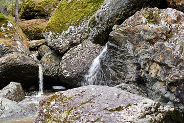 A mountain river waterfall flows through a river bed through huge boulders with moss