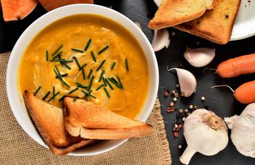 Bowl of sweet potatoes cream soup with fresh chive, roasted toast bread, pepper, carrot and garlic slate slab. View from above