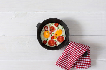 Fresh delicious breakfast of eggs and tomatoes with spices and red pepper, frying pan with fried eggs on white wooden background, top view, copy space