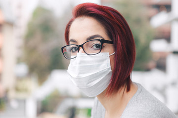 Red-haired young woman wearing glasses and face mask looking at the camera, confined to her home during the coronavirus pandemic
