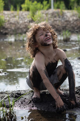 A young boy is playing in the mud, he is very dirty, messy and having a great time. The boy is laughing, happy, the kid feels freedom and free. He is outside, free range child. He is at summer camp