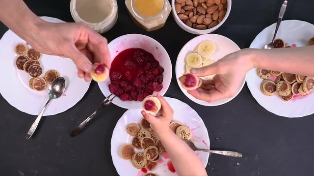 Top view family eating homemade breakfast, clink with mini sandwiches. Tiny pancake cereal, sweet 
spreads, nuts, honey and fresh raspberries. Parents and child eat trendy quarantine food.