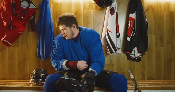 Portrait of angry Caucasian male hockey player in blue uniform sitting on bench in dressing room and looking at camera sadly. Dissapointed sportsman in change room after loose.