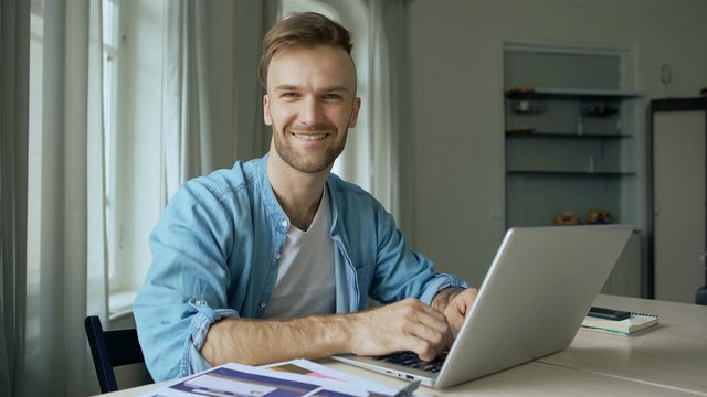 Medium side view shot of young man sitting at desk at home and typing on laptop computer, then smiling, turning head and looking at camera