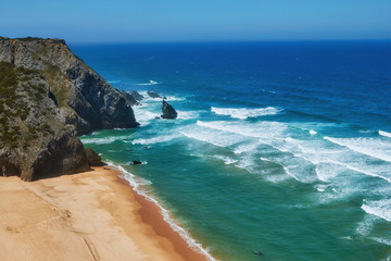 Rocky shore of the Atlantic Ocean. View of high cliffs, foamy waves and sandy beach. Sunny summer seascape. Portuguese riviera. In the area of Cabo da Roca. Portugal.