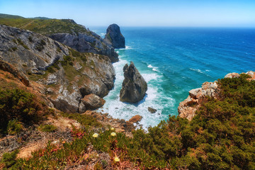 Rocky shore of the Atlantic Ocean. View of high cliffs, foamy waves and sandy beach. Sunny summer seascape. Portuguese riviera. In the area of Cabo da Roca. Portugal.