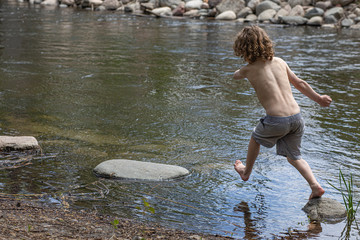 A young boy without his shirt is jumping over rocks in a pond. He is playing in the water, in the summer at camp. Leaping across stones in the creek, pond, lake. Summer vacation, camp. Fun in sun