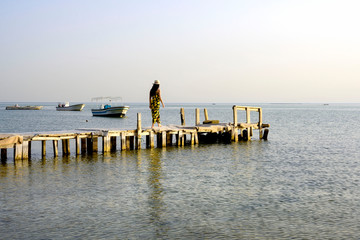 Wooden old pier on calm sea water waves with woman wearing dress and hat walking on over sky and boats parking in the background, Bahrain.