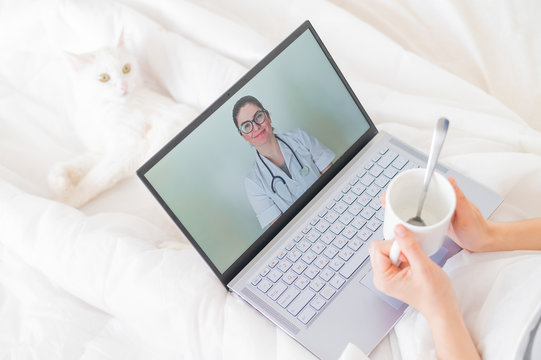 A Faceless Woman Drinks An Antipyretic And Watches A Video From A Doctor Recommendation. Online Doctor Consultation On A Webcam Without Leaving Your Home. The Cat Lies In Bed With A Sick Housewife.