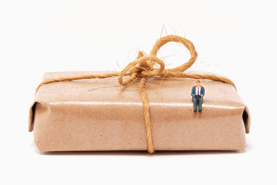 A businessman working on a laptop sits on a classic isolated brown paper wrapped parcel tied with brown parcel string in a bow- Tiny People Working from Home Series