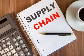 The inscription SUPPLY CHAIN is written in a diary on a wooden table near a calculator and a cup of coffee. Business concept