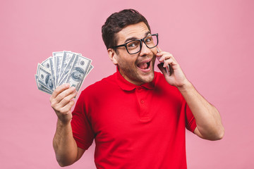 Excited man in casual t-shirt holding lots of money in dollar currencys and cell phone in hands isolated over pink background.