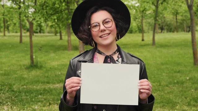 Young hipster woman in a hat and glasses holds a white blank placard in her hands message. Empty area for promo offer. Green park and nature.