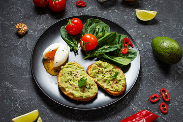Healthy toast with avocado mexican sauce guacamole, boiled egg, cherry tomatoes and salad on black stone background.