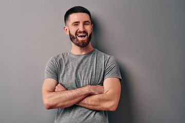 Portrait of a handsome young man leaning against a gray wall crossed his arms and winks and smiles.