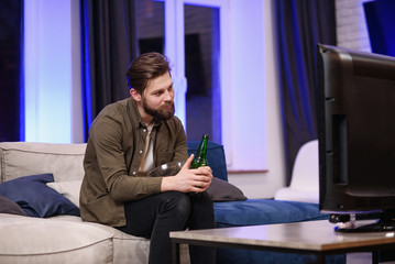 A young man sitting on the sofa in the living room is shocked by a movie on the TV at night.