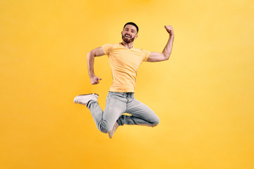 Fototapeta na wymiar Let's dance in the jump! Young man wearing a yellow t-shirt and light jeans jumping against a background of yellow wall showing strong hands.