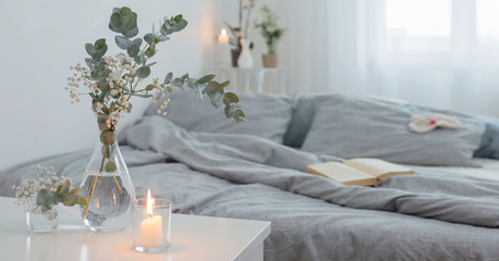 burning candles and eucalyptus in glass vase in white bedroom