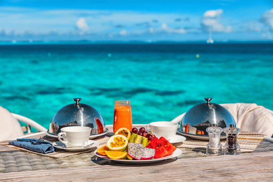 Vacation breakfast at luxury hotel room ocean view. Romantic honeymoon travel holiday in Maldives or Tahiti. Presentation of two plates, fruits, coffee, juice.
