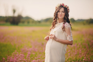 Fototapeta na wymiar Pregnant happy young woman in floral wreath stand on grass in the outdoor in the field of pink flowers background with. Close up.