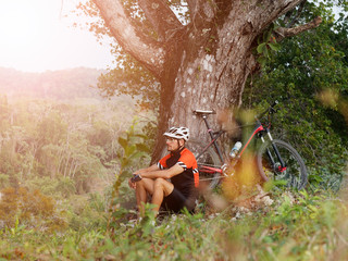 Man riding a mountain bike in the forest. He resting, making pause sits under tree.
