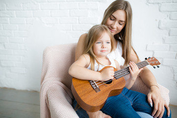 Young mother teaches daughter sitting on her lap on an armchair playing ukulele in the hall