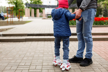 Dad teaches his little daughter to ride four-wheeled roller skates. The father supports the child by the hands, feet of the father and the child in skates. Focus on the rollers. Father's day, family 