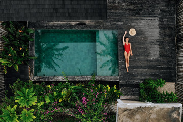 Top view of happy Woman in red swimsuit and straw hat sunbathe near private pool with flowers and greenery around, Bali. Tropical background and travel concept.