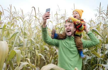 Father carries little son in his shoulders and make selfie while walking across yellow autumn corn...