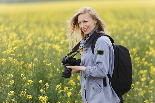 Charming lady in denim shirt standing at green field with big backpack and digital camera. Female photographer with blond hair taking pictures of beautiful spring nature