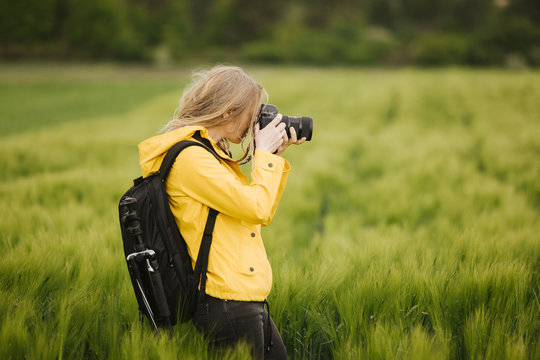 Side view of concentrated woman in yellow jacket taking photos of beautiful wheat field. Female photographer with black backpack enjoying working process outdoors.