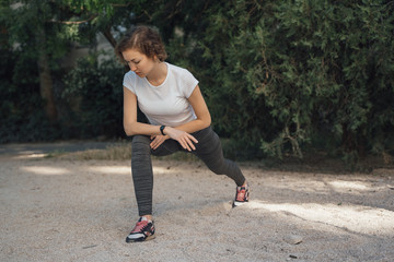 A girl with blond blonde curly hair in a white top and gray leggings does sports exercises outdoors