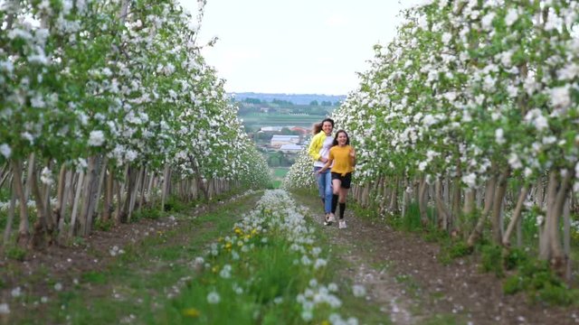 Mom and her daughters in casual clothes run across the green lawn in blooming apple blossom garden. Family concept