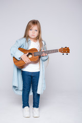 Fototapeta premium Full-length portrait of a little girl musician holding a ukulele in her hands on a light background. Shooting for a music school