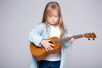A blond girl a child of European appearance plays a small guitar on a gray background