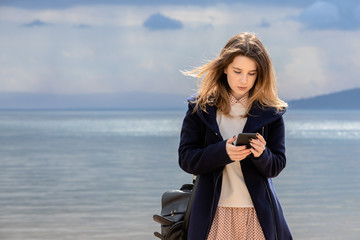 A young adult girl with a smartphone stands on the seashore against a dark cloudy sky.