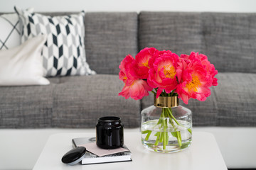 Bouquet of the pink peonies and book with black candle on the white table and sofa with pillows on the background. Flowers in vase. Stylish interior.