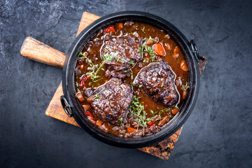Traditional German braised beef cheeks in brown red wine sauce with carrots and onion offered as top view in a modern design stewpot on an old rustic board - obrazy, fototapety, plakaty
