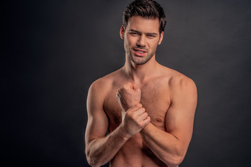 Fototapeta premium Handsome young bearded man isolated. Cropped image of topless muscular man is standing on gray background. Man holding his wrist. Experiencing wrist pain.