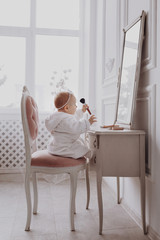 A cute little girl is holding a makeup brush and having fun at home. baby Girl is sitting on the chair near the classic mirror indoors . Kid's fashion. Little girl fashionista.