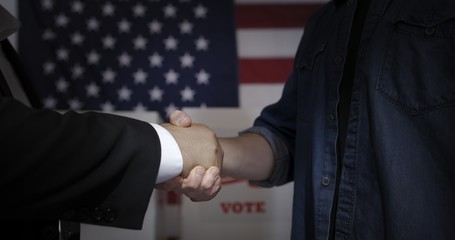 CU detail of hands as anonymous man in shirt and jacket hands shaking hands with an unseen young man in front of voting booths and American flag.