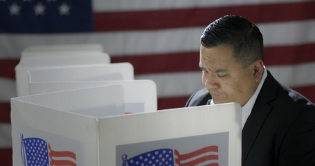 MS Hispanic man in polling station, voting in a booth with US flag in background. Serious...