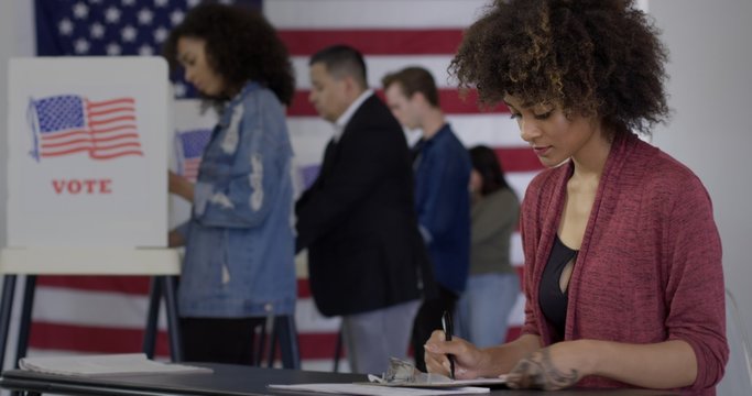 MS Young mixed-race woman staffing desk at polling station with various voters in background, US flag on wall behind them.