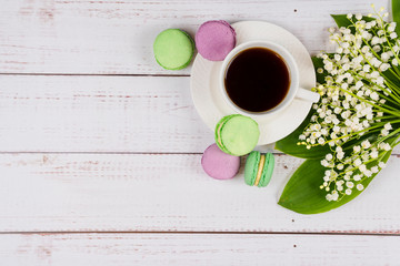 Obraz na płótnie Canvas Colorful macaroons, a cup of coffee and lilies of the valley on wooden background, close-up flat lay.