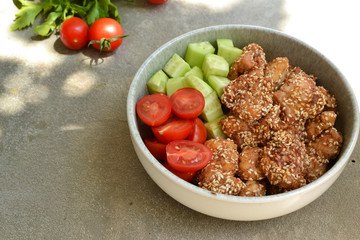 Tasty lunch in a gray bowl on a concrete background. Nuggets with sesame seeds. Cucumbers and tomatoes. Salad and chicken. Free space for text