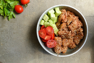 Tasty lunch in a gray bowl on a concrete background. Nuggets with sesame seeds. Cucumbers and tomatoes. Salad and chicken. Free space for text Top view