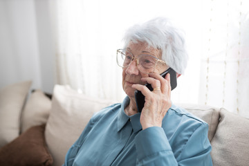 Sad and lonely senior gray-haired Caucasian woman sitting on a sofa in brightly lit living room and talking on the phone