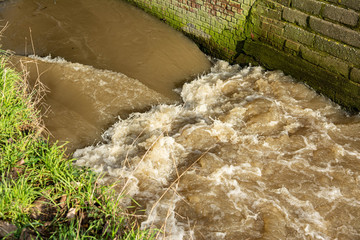 Little river canalized between walls. A difference in level causes swirling and foaming.The water is brownish, on one side a moss green brick wall, on the other a grassy bank.