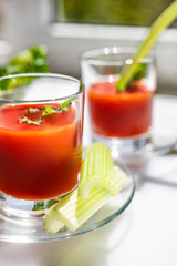 Two glasses of tomato juice with parsley and celery decorations, stand near the window, morning sunlight shines, shallow depth of field, selective focus. Natural drinks concept.