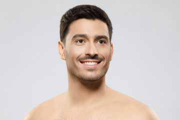 Close-up of young handsome male with short brown hair, bristle and naked shoulders, looking aside with hopeful smile, isolated on gray background